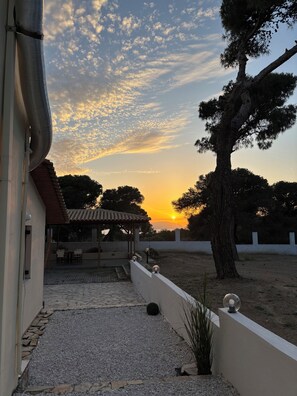 Um edifício com varanda coberta, uma árvore e um pôr do sol ao fundo.