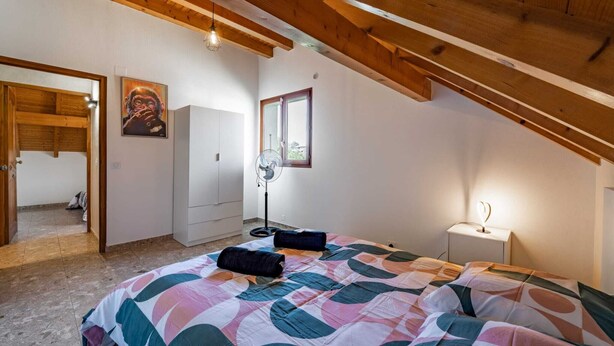 Una camera da letto con soffitto spiovente, un letto con copriletto colorato, una ventola da terra, una lampada da comodino e un quadro appeso al muro.
