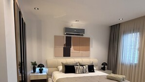 A modern bedroom with a large bed, a bedside table with a lamp, a wall-mounted air conditioner, and a window with curtains. - Luxury Renovated New Villa in the heart of Kalkan (ANTALYA)