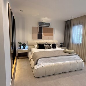 Ein modernes Schlafzimmer mit einem großen Bett, einem Nachttisch mit Lampe, einer Wand-Klimaanlage und einem Fenster mit Vorhängen.