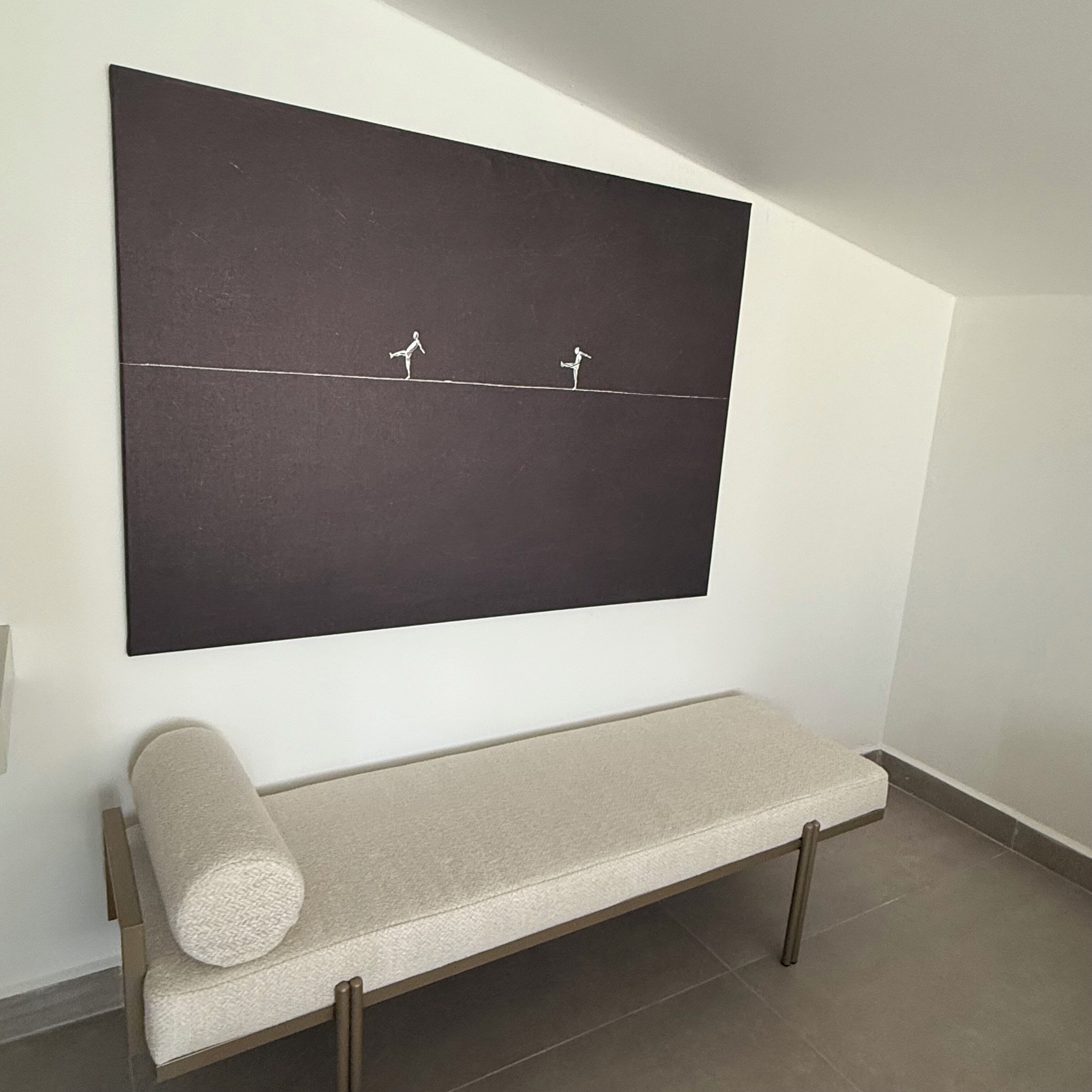 A room with a large black artwork on the wall, a bench with a cushion, and a tiled floor.