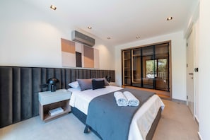A modern bedroom with a large bed, a bedside table with a lamp, a wall-mounted air conditioner, and a sliding glass door leading to a balcony. - Luxury Renovated New Villa in the heart of Kalkan (ANTALYA)