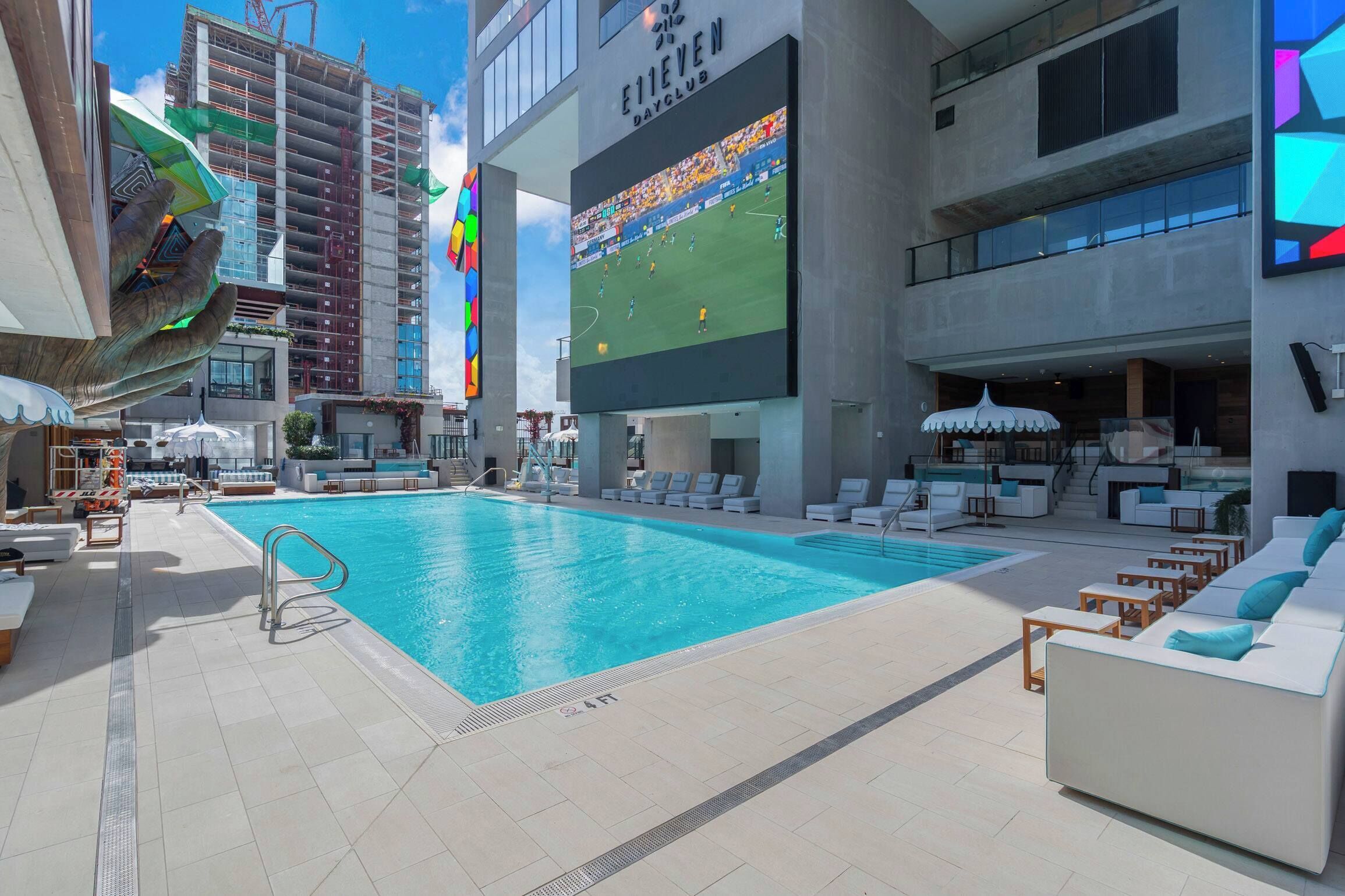 A rooftop pool area with lounge chairs and a large screen displaying a sports event.