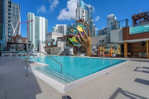 Ein Schwimmbecken mit einer großen Hand-Skulptur, umgeben von modernen Gebäuden und Baukränen.