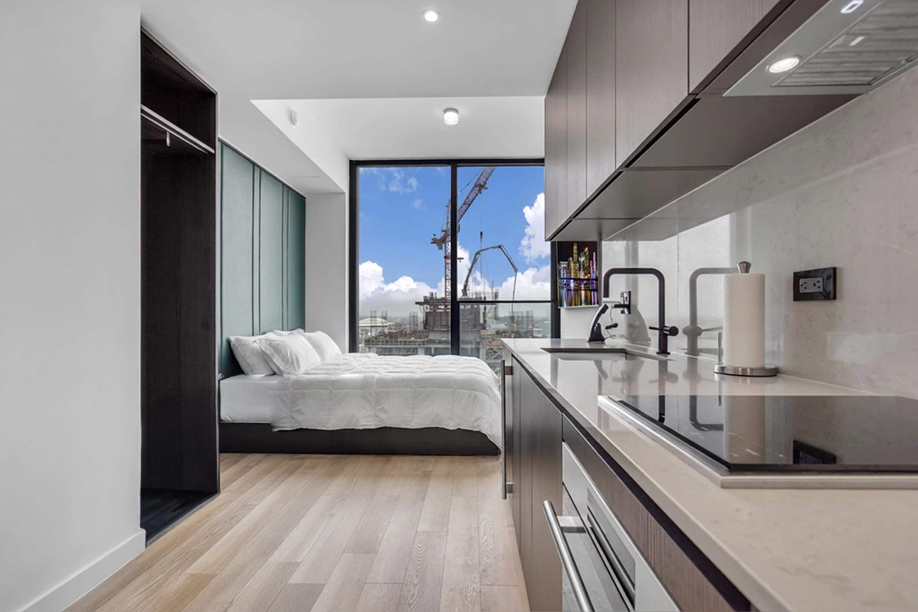 A modern bedroom with a bed, a built-in wardrobe, a large window with a city view, and a kitchenette.
