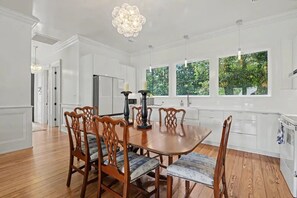 A dining room with a wooden table and chairs, white cabinetry, and large windows with a view of greenery. - Brand New Beautiful House on the Water in Timber Ridge (Pass Christian)