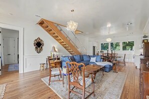 A spacious living room with a wooden staircase, a blue sofa set, and a dining area with wooden furniture. - Brand New Beautiful House on the Water in Timber Ridge (Pass Christian)