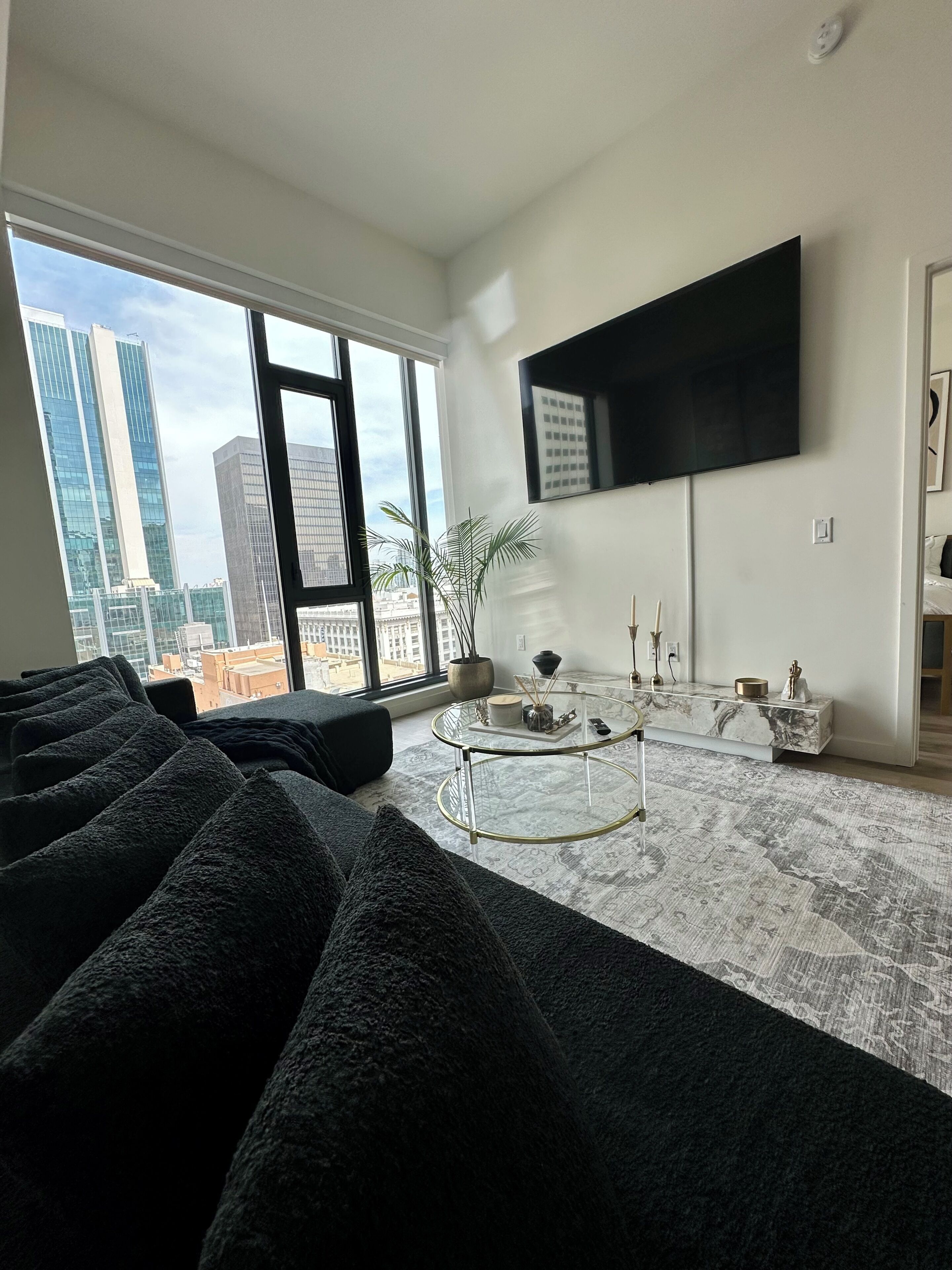 A modern living room with a large window, a flat-screen TV mounted on the wall, a glass coffee table, and a patterned rug.