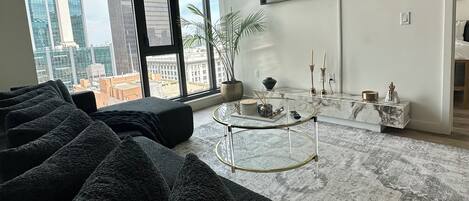 A modern living room with a large window, a flat-screen TV mounted on the wall, a glass coffee table, and a patterned rug.