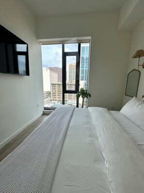 A hotel room with a large bed, a flat-screen TV mounted on the wall, and a view of the cityscape through the window. - Cozy 1-bedroom apartment with Luxury Amenities in Downtown San Diego (San Diego)