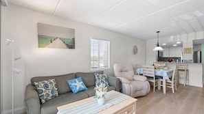 Ein modernes Wohnzimmer mit grauem Sofa, gemusterten Kissen, einem Couchtisch mit einer Vase, einem Essbereich mit Holzkästen und einer Küche im Hintergrund.