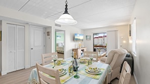 A dining area with a table set for four, a television mounted on the wall, and a view of the outside through a window. - Coastal St. Augustine Beach Condo | Partial Ocean View | Resort Amenities | Steps to the Beach | Sleeps 6 (St. Augustine)