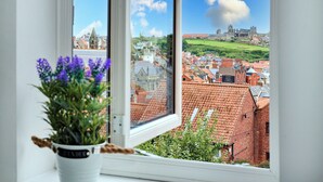 A view through a window with a potted plant in the foreground, overlooking a town with red-roofed buildings and a distant castle-like structure. - Holiday Home Near Centre of Whitby (Whitby)