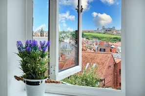 Blick durch ein Fenster mit einem Blumentopf im Vordergrund auf eine Stadt mit roten Dächern und einer weit entfernten, schlossähnlichen Struktur.