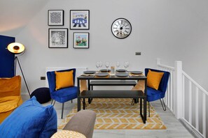 Un coin repas avec une table dressée pour quatre personnes, deux chaises bleues avec des coussins jaunes, une horloge noire au mur et des œuvres d’art encadrées au-dessus de la table.