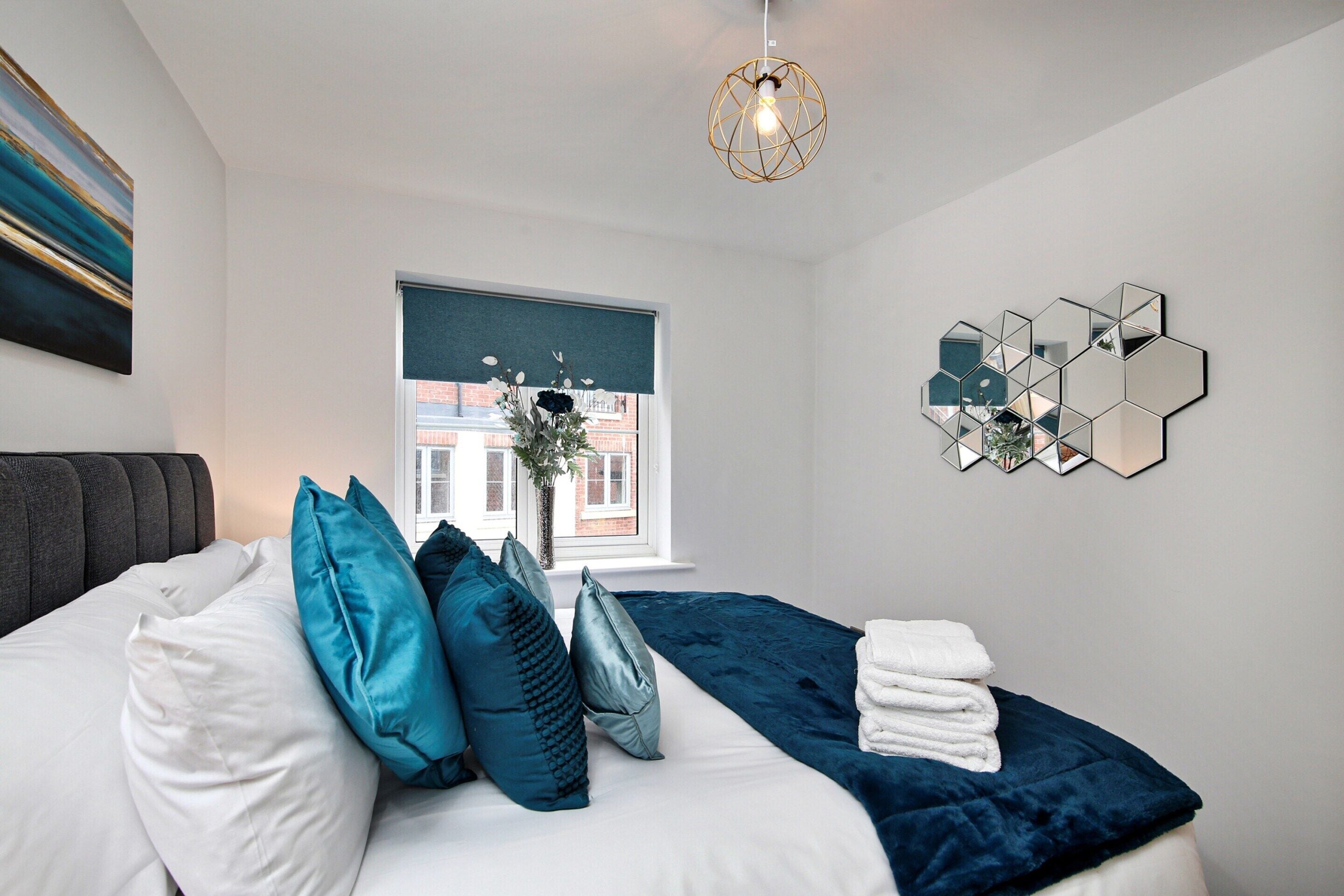 A modern bedroom with a bed, pillows, a window with a view, a hanging light fixture, and a wall-mounted mirror.