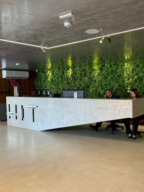 A reception desk with a 'WELCOME HIT' sign, a green wall with plants, and a ceiling with recessed lighting. - HIT Cabo Branco (João Pessoa)