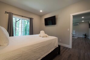 A bedroom with a large bed, a flat-screen TV mounted on the wall, and a view of a forest through the window.