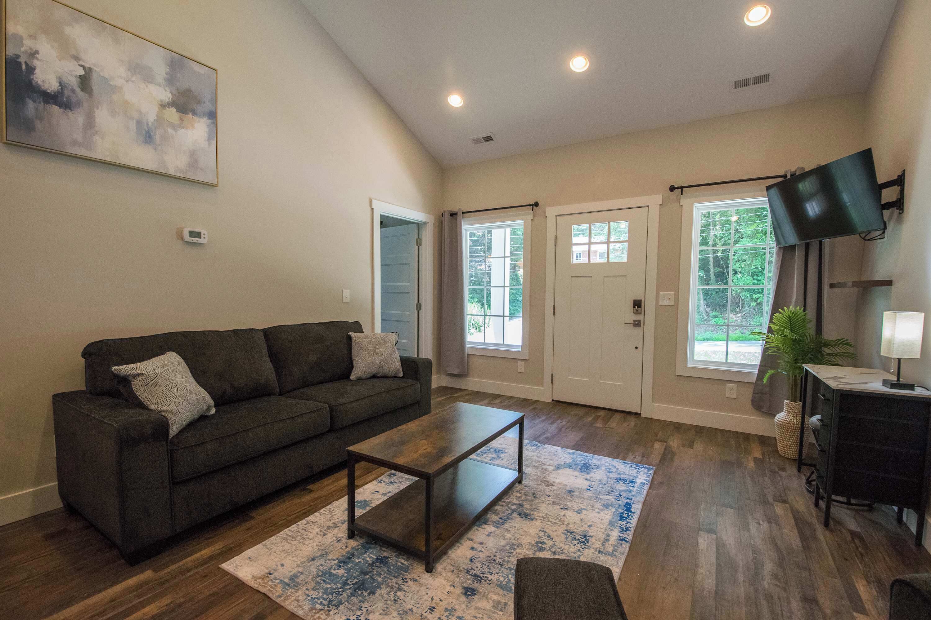 A living room with a dark grey sofa, a coffee table, and a patterned rug. There are two doors leading outside, a flat-screen TV mounted on the wall, and a small table with a lamp.