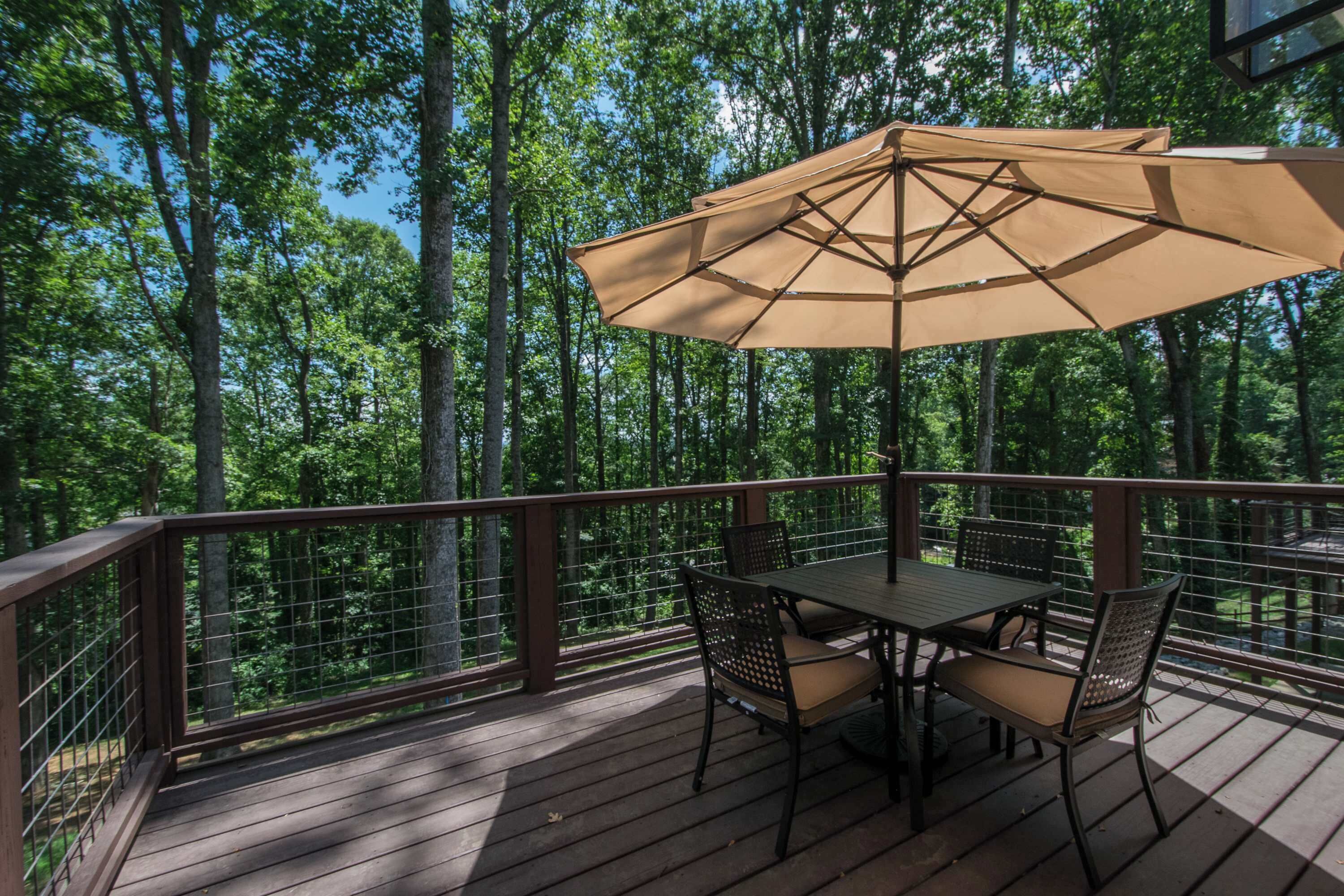 Una terraza de madera con mesa y sillas, rodeada por una barandilla de metal, con vistas a un bosque.