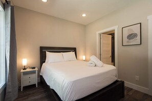 Une chambre d’hôtel avec un lit simple, des tables de chevet, une petite table, une lampe, un tableau encadré au mur et une porte donnant sur une autre pièce.