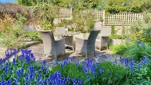 An outdoor patio with wicker furniture and a stone floor, surrounded by lush greenery and flowering plants. - Cherry Tree Cottage - sleeps 6 guests  in 3 bedrooms (St Minver)