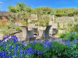 Eine Terrasse im Freien mit Korbmöbeln und Steinboden, umgeben von üppiger Vegetation und blühenden Pflanzen.