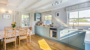 A modern kitchen with a dining area, featuring wooden furniture, a built-in oven, and a large window providing natural light. - Cherry Tree Cottage - sleeps 6 guests  in 3 bedrooms (St Minver)