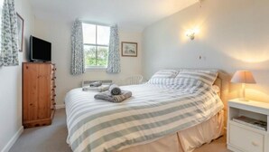 A bedroom with a bed, a TV, a window with curtains, a bedside table with a lamp, and a wooden wardrobe. - Cherry Tree Cottage - sleeps 6 guests  in 3 bedrooms (St Minver)