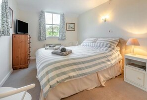 Ein Schlafzimmer mit Bett, Fernseher, Fenster mit Vorhängen, Nachttisch mit Lampe und einem Holzschrank.
