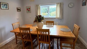 A dining area with a wooden table and chairs, a vase with flowers, a clock on the wall, and a window with curtains. - Broad Park-Sleeps 11 in 6 Bedrooms (Rock)
