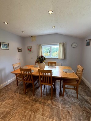 A dining area with a wooden table and chairs, a vase with flowers, a clock on the wall, and a window with curtains. - Broad Park-Sleeps 11 in 6 Bedrooms (Rock)
