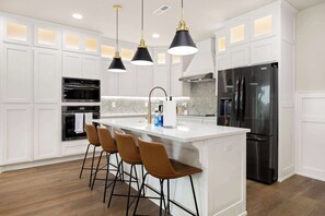 Cocina moderna con armarios blancos, una isla central con taburetes de bar, electrodomésticos de acero inoxidable e iluminación colgante.