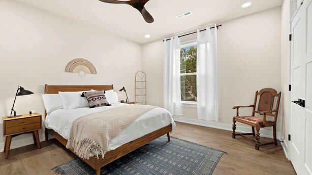 Un dormitorio con cama, mesita de noche, silla, ventilador de techo y ventana con cortinas.
