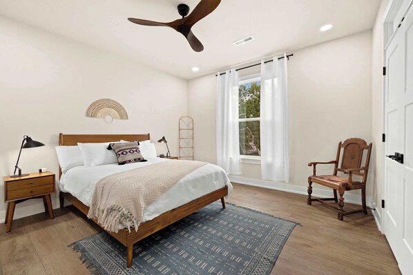 A bedroom with a bed, a nightstand, a chair, a ceiling fan, and a window with curtains.