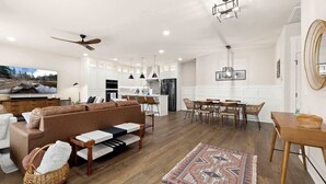 Una sala moderna con un sofá marrón, un comedor con sillas de madera, una cocina con armarios blancos y un ventilador de techo.