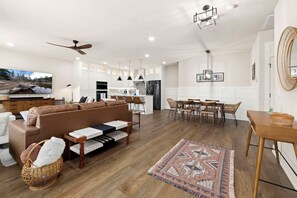 A modern living room with a brown sofa, a dining area with wooden chairs, a kitchen with white cabinets, and a ceiling fan.