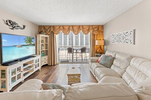 A living room with a television, sofa, and a view of a balcony with outdoor furniture. - S Hampton 706 2 Br Oceanview in Kingston, Pool (Myrtle Beach)