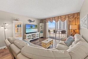 Ein Wohnzimmer mit einer Couch, einem Fernseher und Blick auf den Strand durch große Fenster.