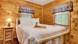 A wooden cabin bedroom with a bed, bedside table, lamp, and windows with curtains. - Dream Chaser by Avantstay Hot Tub + Arcade + Porch Swing Mountain Views, Minutes to the Parkway (Sevierville)