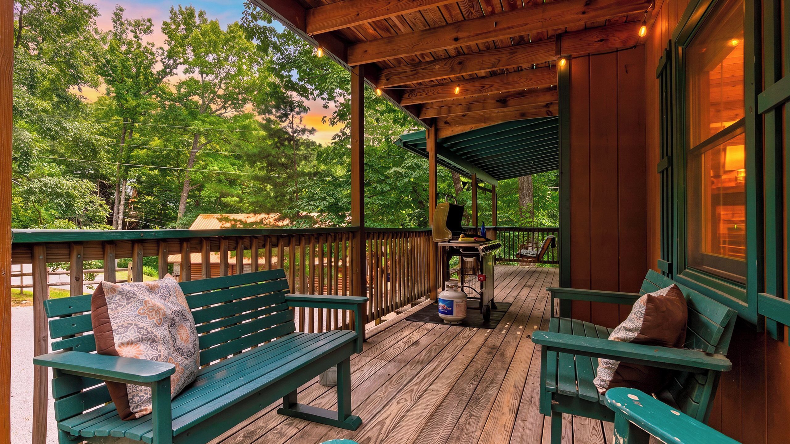 A wooden deck with green benches and a grill, surrounded by trees.