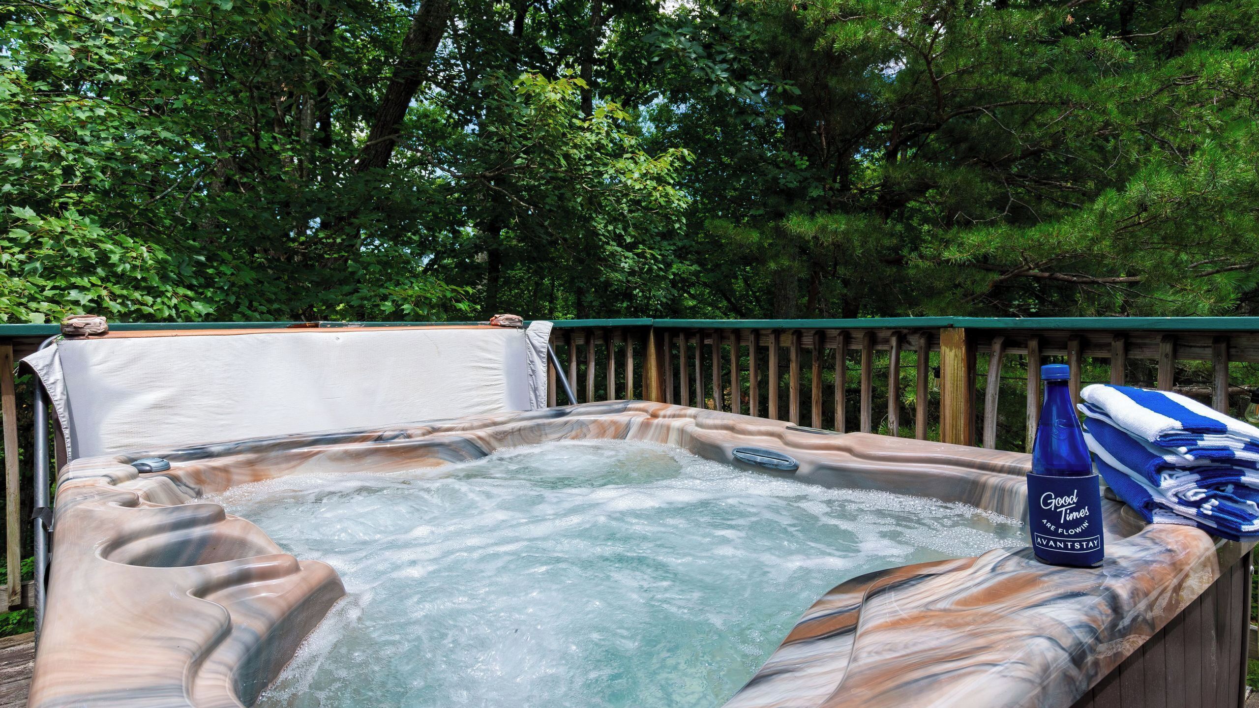 A hot tub surrounded by a wooden deck with a green railing, set in a wooded area.