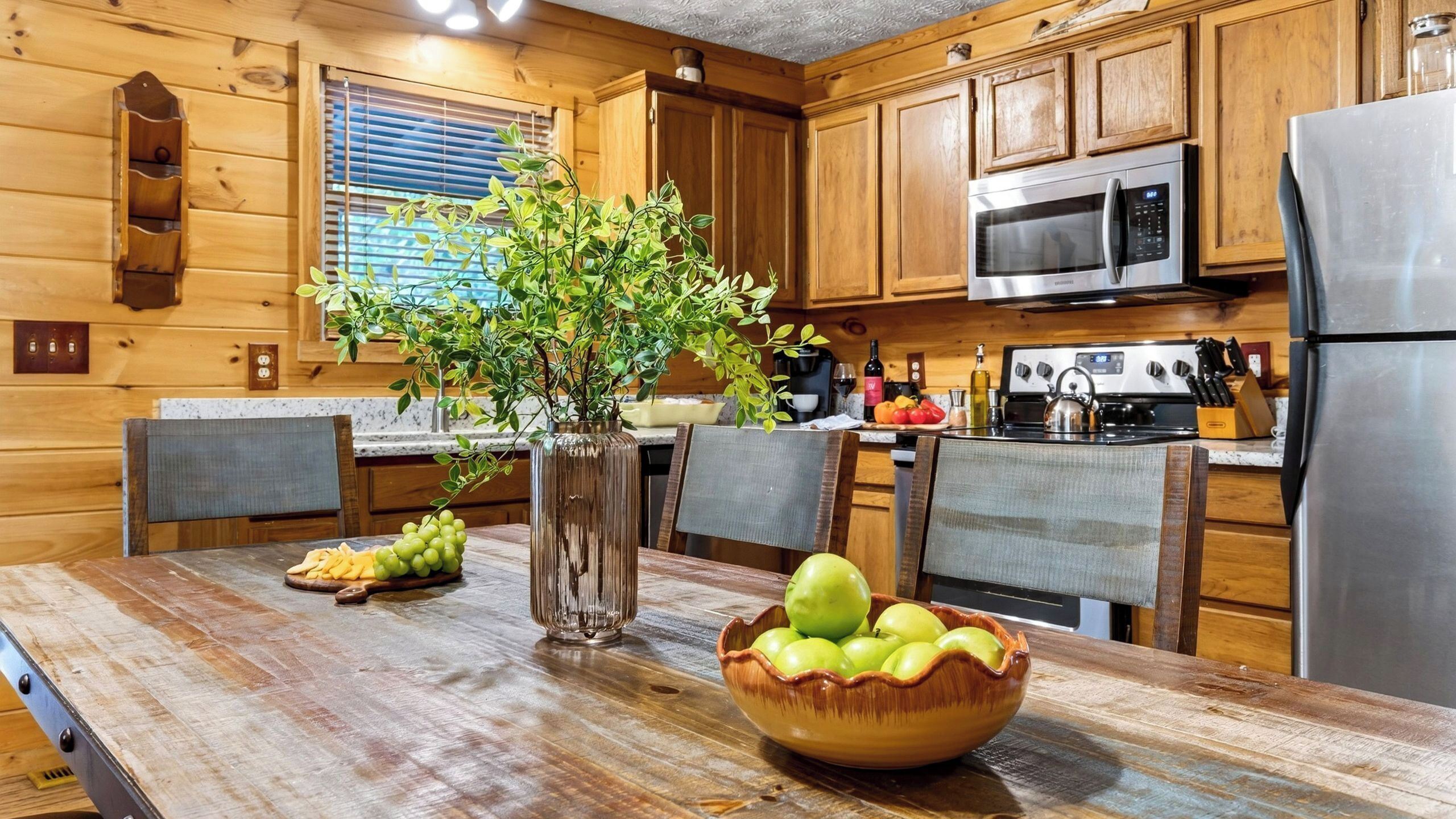 A wooden cabin kitchen with a dining table, a vase with green plants, a bowl of green apples, and modern appliances.