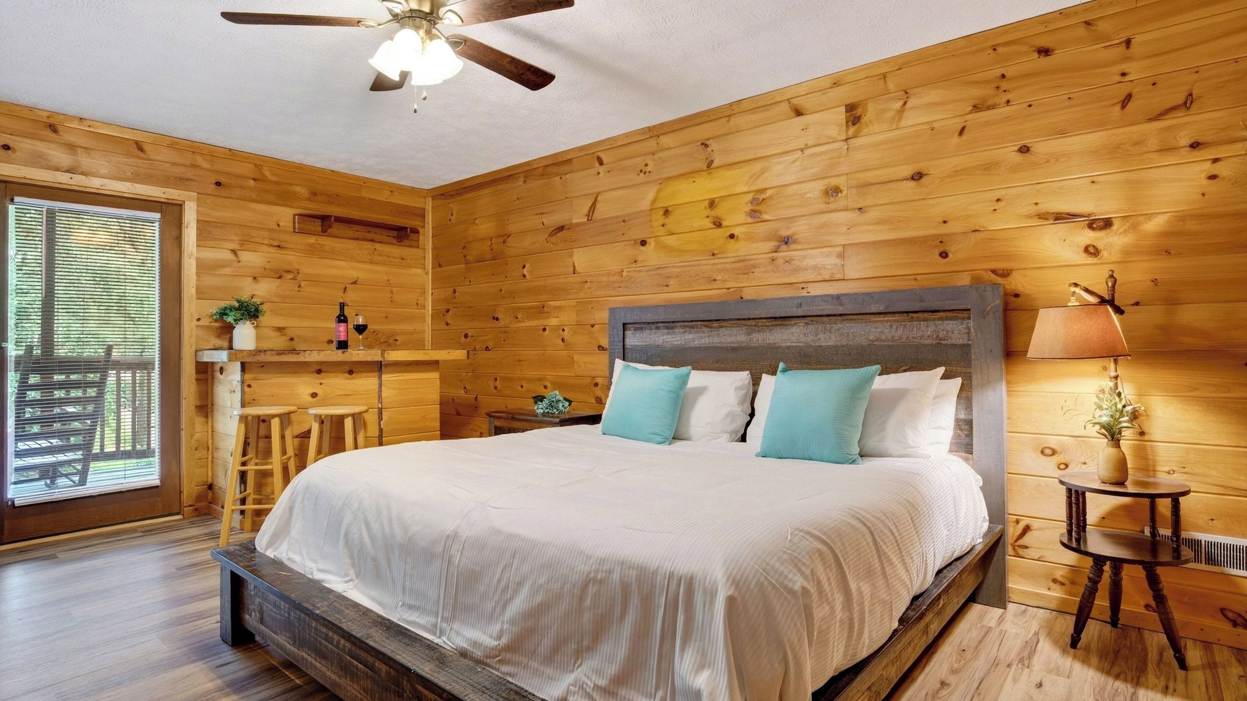 A wooden-paneled bedroom with a bed, a ceiling fan, a nightstand, a lamp, and a window with blinds.