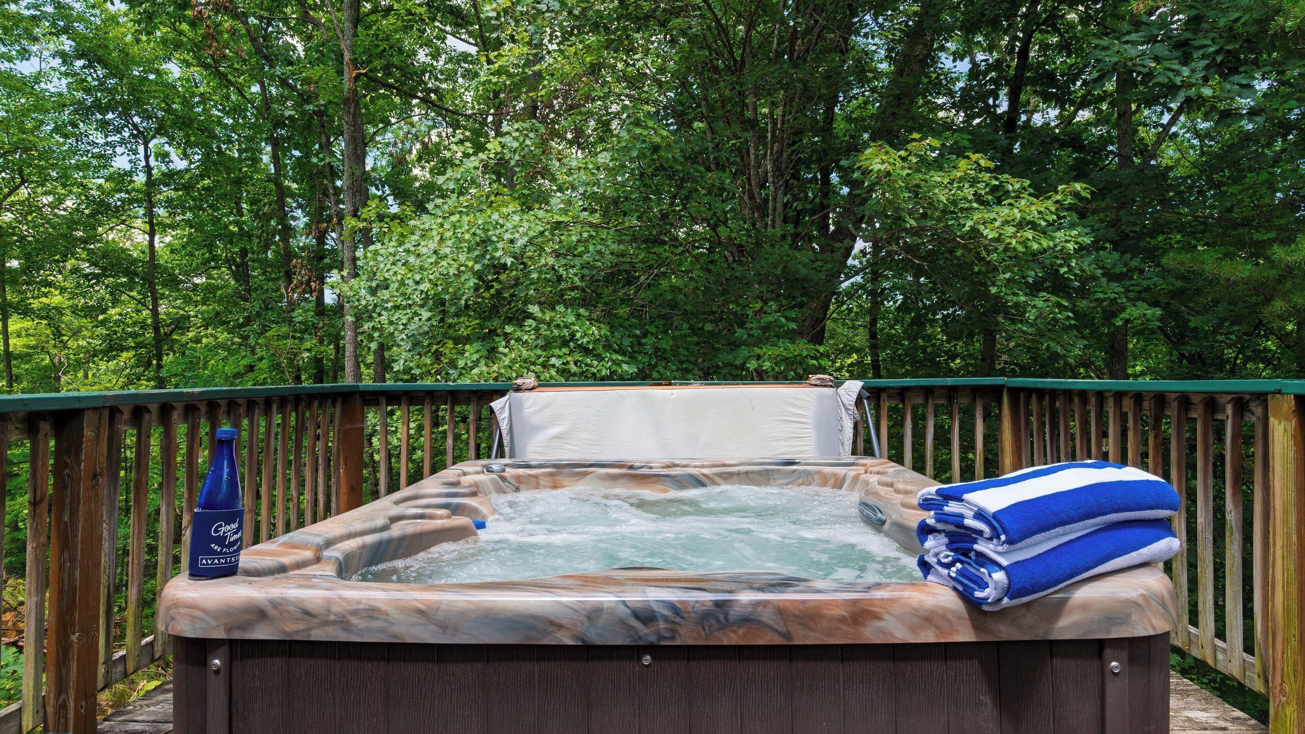 A hot tub outdoors surrounded by a wooden deck and dense forest.
