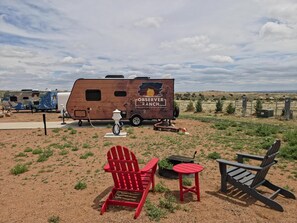 En campingplads med en campingvogn i træ, røde og sorte Adirondack-stole og et lille bord.