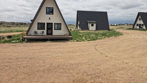 Um edifício triangular com varanda e duas cadeiras, uma estrutura menor anexa, e um caminho de terra que leva a ele.