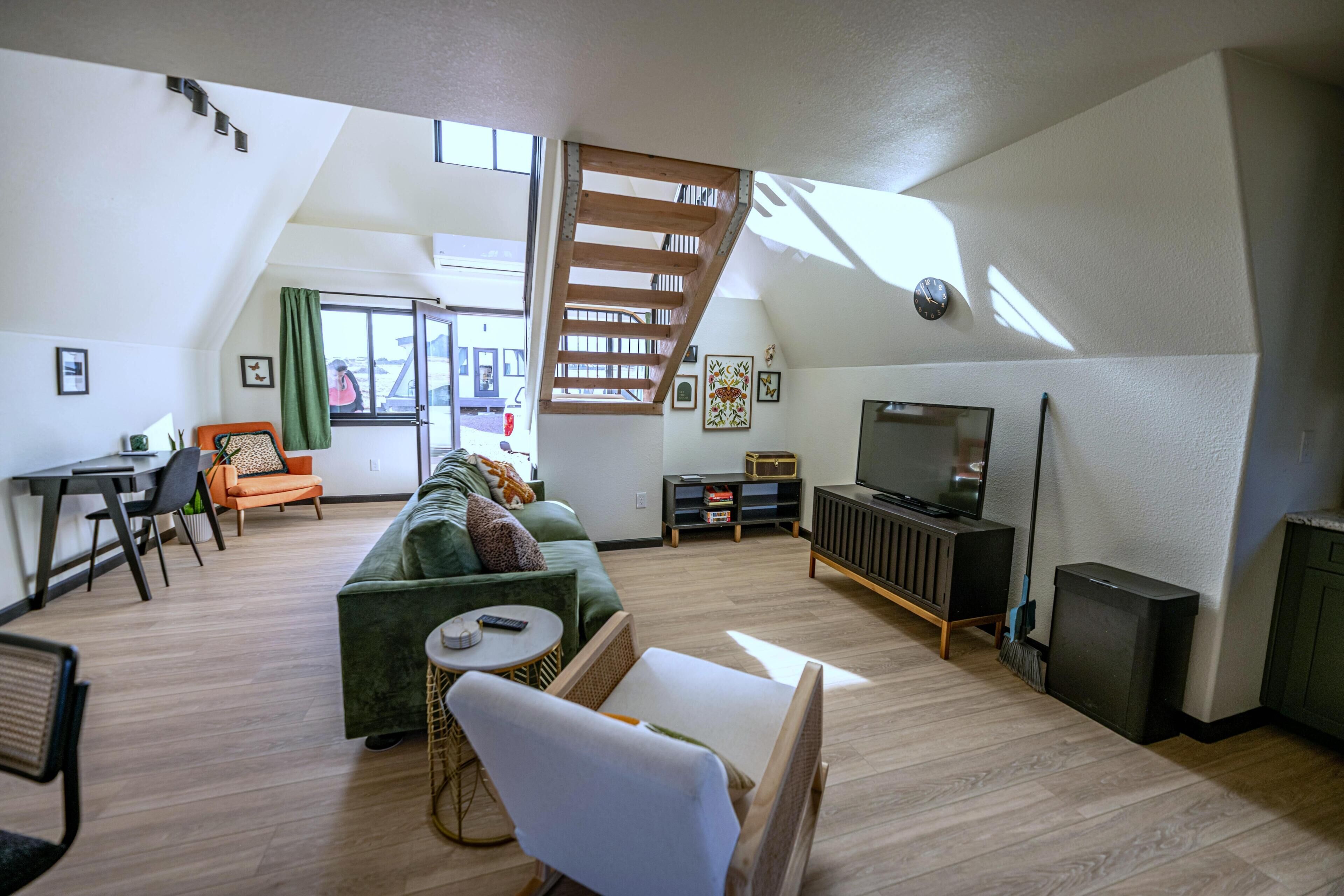 A spacious living room with a green sofa, a white armchair, and a TV on a stand. A wooden staircase leads to an upper level.