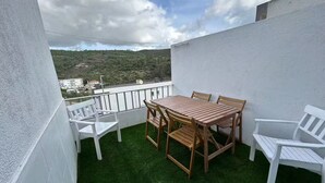 En balkon med et træbord og stole, omgivet af hvide vægge og udsigt over et bølgende landskab.