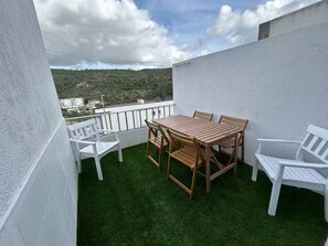 Ein Balkon mit Holztisch und -stühlen, umgeben von weißen Wänden und Blick auf eine hügelige Landschaft.